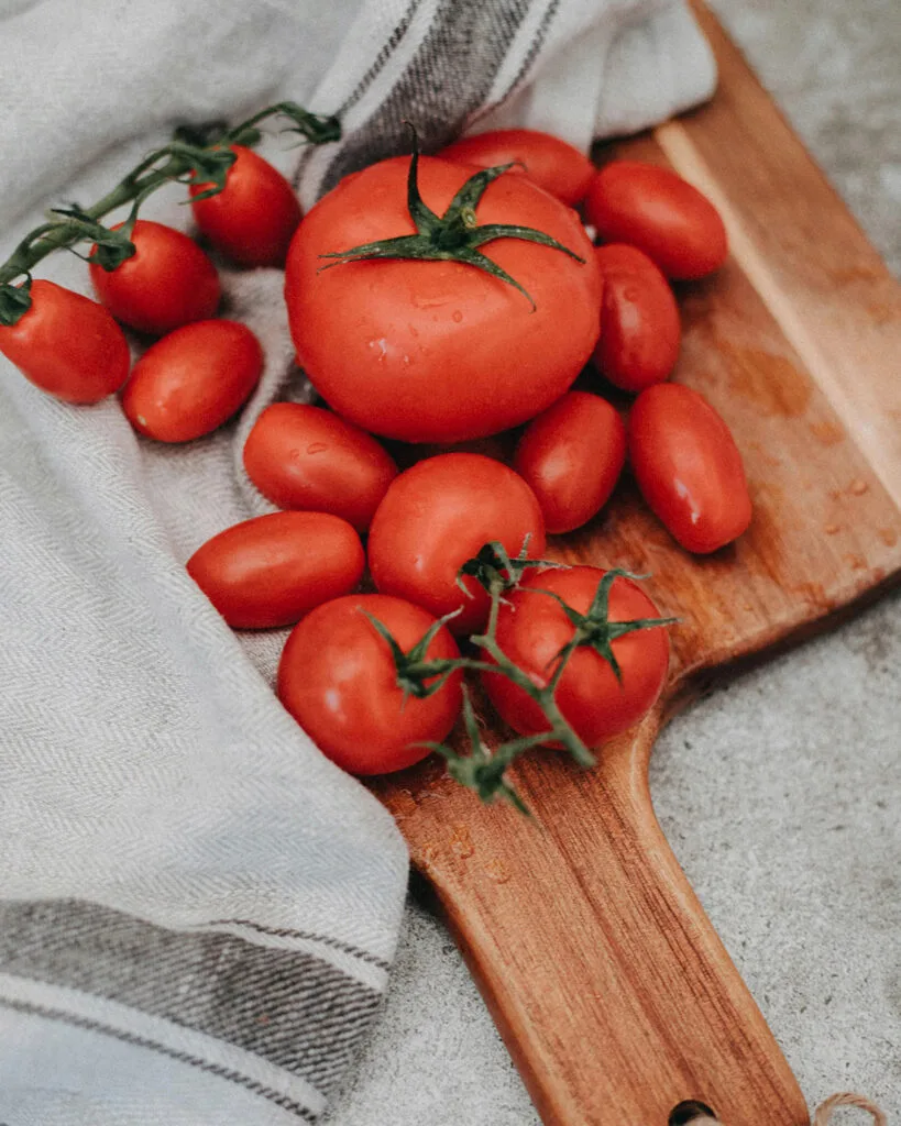 A variety of red tomatoes of different sizes and shapes on a wooden cutting board with a white cloth.